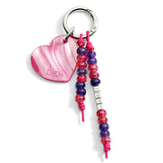 Charm a forma di cuore in bioresina vegetale color rosa fucsia e viola, con dettagli in acciaio lucido, appeso a una borsa elegante.